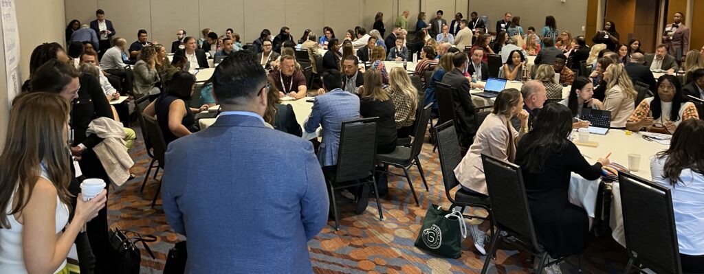 A diverse audience is discussing at round tables. The room is full with more people standing at the sides and in the front.