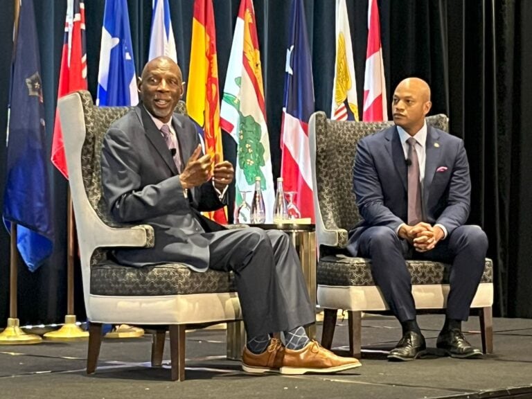 Geoffrey Canada with Maryland Governor Wes Moore seated on a stage with state, province and territorial flags in the background.