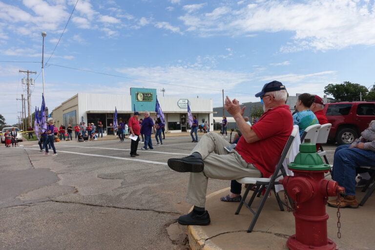 A senior man with white hair is seated on a folding chair on the curb in a small downtown. He's applauding for a marching flag group with diverse kids in a parade.