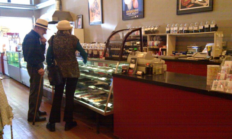 In a quaint local candy store, an older couple both wearing hats and using canes for mobility are browsing for sweet treats.