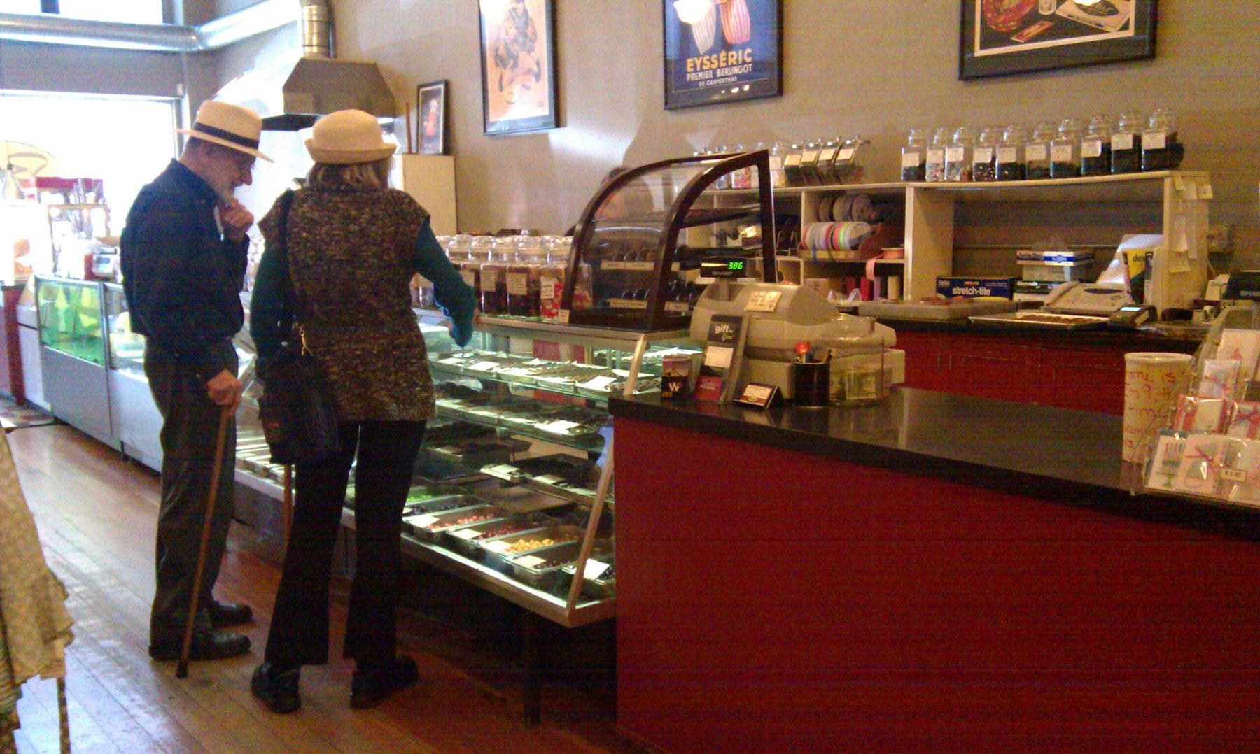 In a quaint local candy store, an older couple both wearing hats and using canes for mobility are browsing for sweet treats.