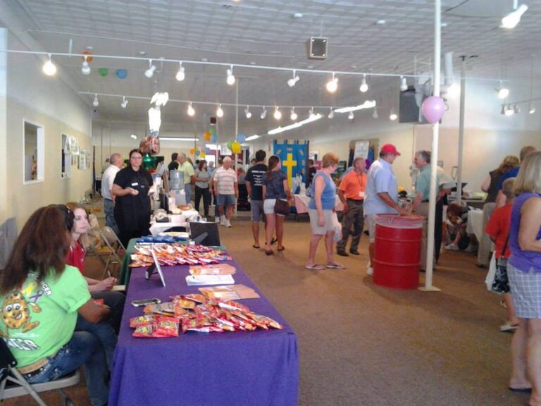 An empty building in a small downtown has been filled with booths for local businesses. A crowd is circulating among the colorful booths and chatting with each other.