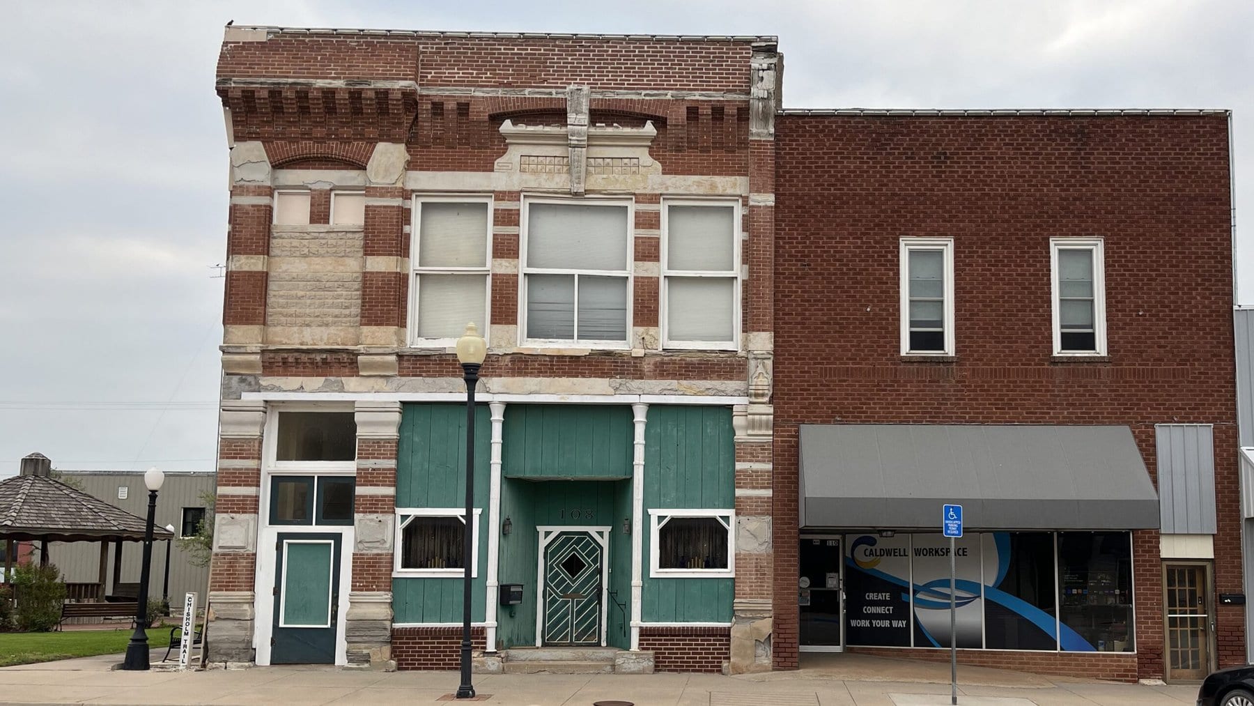 A plain brick building in a small downtown has a window decal that says “Caldwell Workspace: create, connect, work your way” Next door is an ornate brick building from the late 1800s.