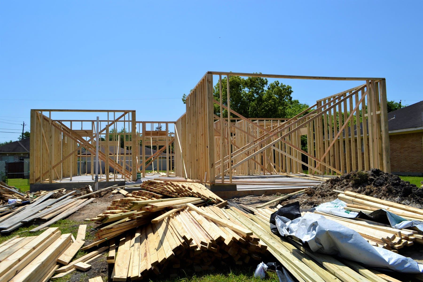 A house is shown in the framing stage, with the stud walls up. A stack of lumber sits in front of it.