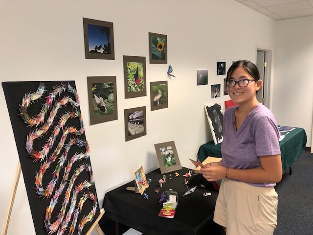 A young person showcasing origami art and photos from a table set in an empty building. 